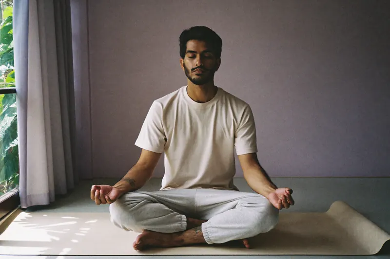 A man meditates cross-legged on a mat by a window, eyes closed, in soft afternoon light