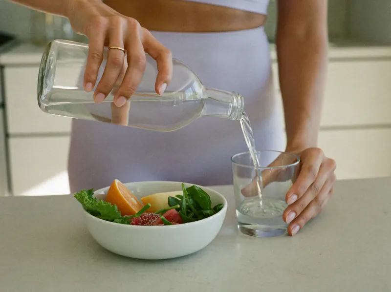 Hands pour a glass of water beside a bowl of salad and fruit on a kitchen counter